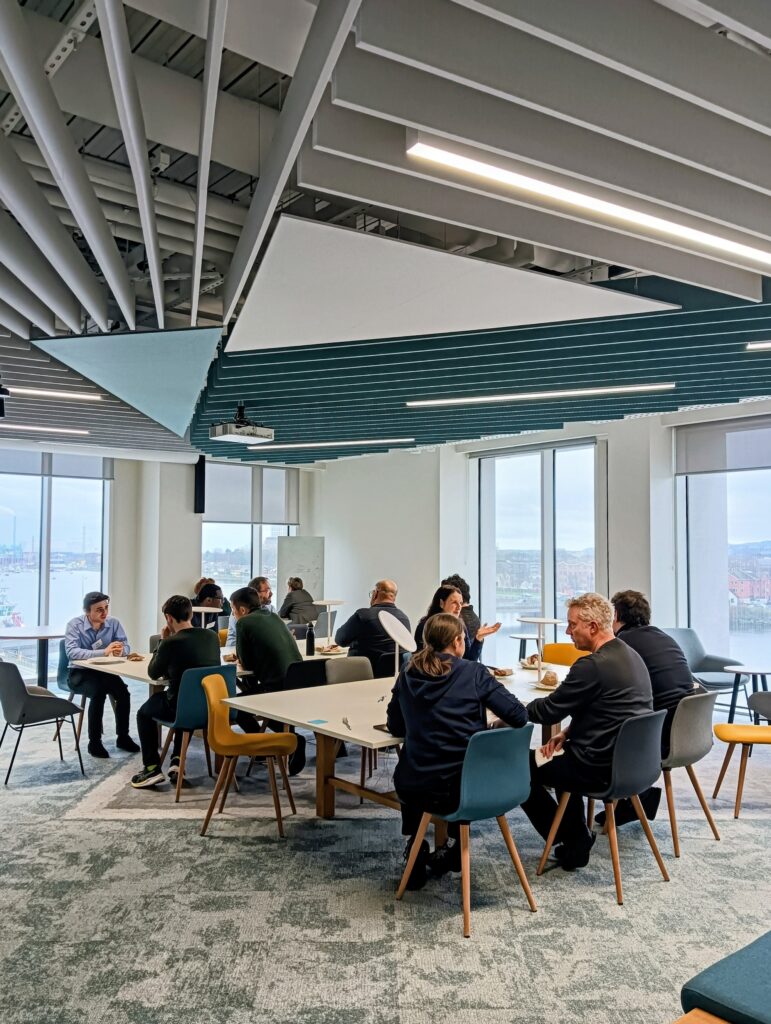 People sit around two large boardroom tables in an office with large glass windows.